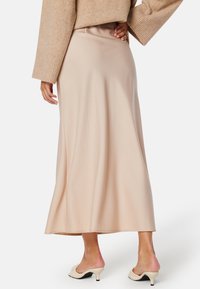 Beige saténová maxi sukně s volným střihem, hladkým povrchem a mírným leskem, spárovaná s bílými otevřenými podpatky.