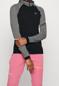 Giacca con zip nera e grigia con cappuccio, maniche grigie testurizzate e corpo nero. Presenta una zip rosa e un logo rosa sulla manica.