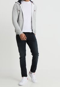 Haut à capuche gris clair avec des accents bleu marine, porté sur un t-shirt blanc. Associé à un jean sombre et des baskets blanches avec des détails rouges.