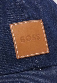 Denim pet met een bruine leren label met het met de hand gestempelde "BOSS" logo, witte stiksel en een gestructureerd ontwerp in diepblauw.