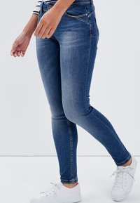 Schmale blaue Jeans aus Denim mit einem ausgewaschenen Muster, fünf Taschen und kontrastierenden Nähten, kombiniert mit weißen Sneakern.