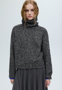 Szürke kötött garbó pulóver texturált felülettel, laza szabással és bordázott szegéllyel. Egy sötétebb szürke szoknya fölött viselve, kék mandzsettákat mutatva.