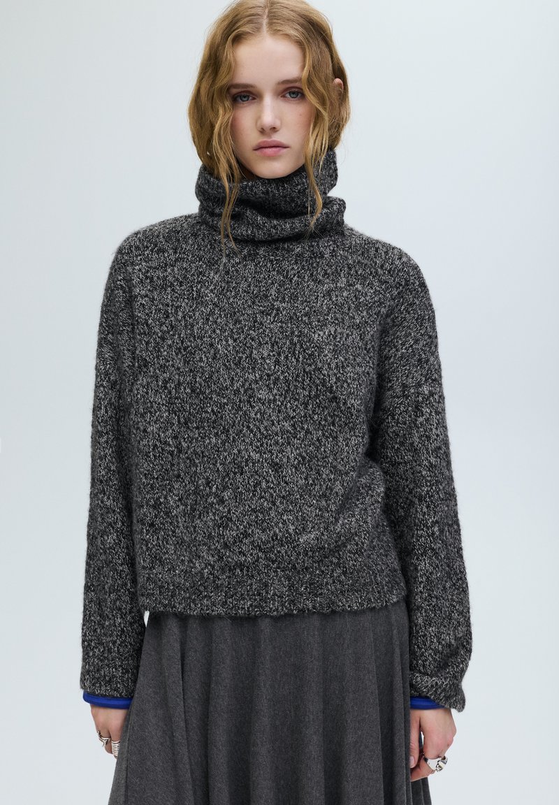 Szürke kötött garbó pulóver texturált felülettel, laza szabással és bordázott szegéllyel. Egy sötétebb szürke szoknya fölött viselve, kék mandzsettákat mutatva.