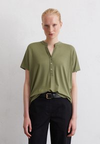 Olijfgroene shirt met korte mouwen, voorzien van een halve knoplijst en een kraag, gecombineerd met zwarte broek en een zwarte riem met een gouden gesp.