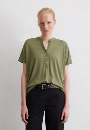 Olijfgroene shirt met korte mouwen, voorzien van een halve knoplijst en een kraag, gecombineerd met zwarte broek en een zwarte riem met een gouden gesp.