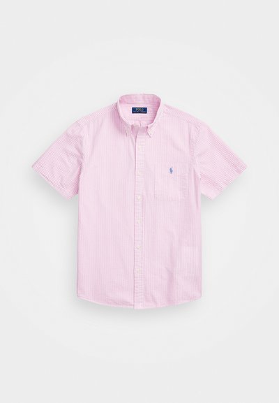 Īsām piedurkņu pogājamā krekla gaiši rozā krāsā ar plānām baltām svītrām. Iekļauj apkakli, priekšējo kabatu un zilu izšūtu logotipu uz krūtīm.