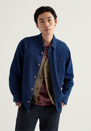 Jauns vīrietis ar melniem matiem, ģērbies vairākos apģērbos: zila džinsa jaka, bēša pogām aiztaisīta veste un sarkanmelna svītraina krekls, rokas kabatās.