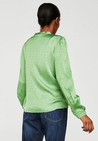 Blusa verde in satin con maniche lunghe, spalle arricciate, texture a pois e polsini arricciati. La vista posteriore mostra un collo rotondo e una vestibilità ampia.