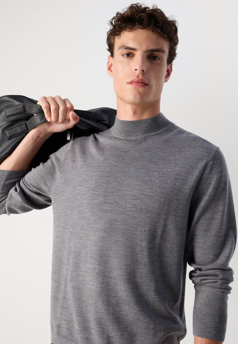 Grauer Strickpullover mit hohem Kragen und gerippten Bündchen, mit einem kleinen gestickten Logo auf der Brust. Der Stoff wirkt glatt und weich.