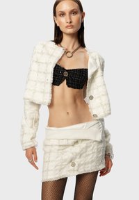 Witte, structuurrijke cropped blazer met zilveren knopen, gelaagd over een zwarte strapless top en een witte minirok. Het model draagt een visnetpanty.
