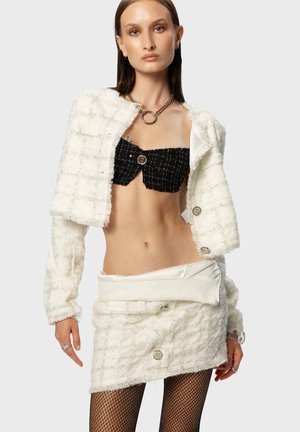 Giacca corta bianca a trama con bottoni argentati indossata sopra un top senza spalline nero e una mini gonna bianca. La modella indossa collant a rete.