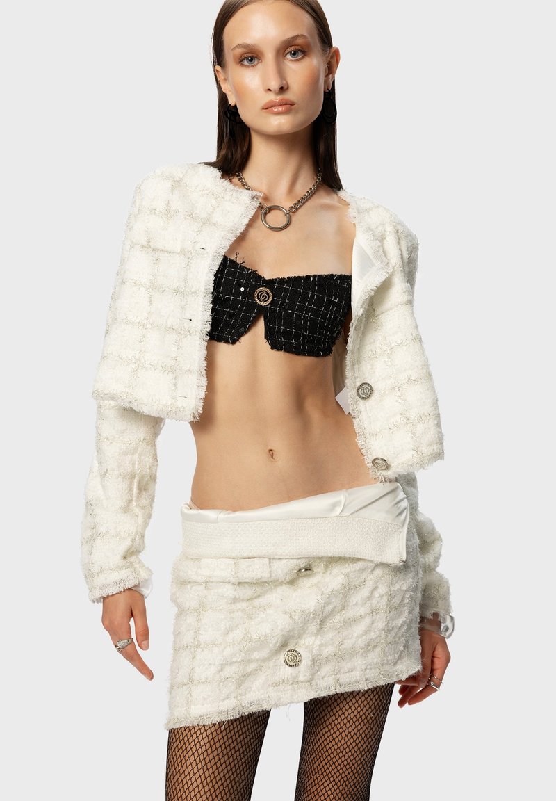 Witte, structuurrijke cropped blazer met zilveren knopen, gelaagd over een zwarte strapless top en een witte minirok. Het model draagt een visnetpanty.