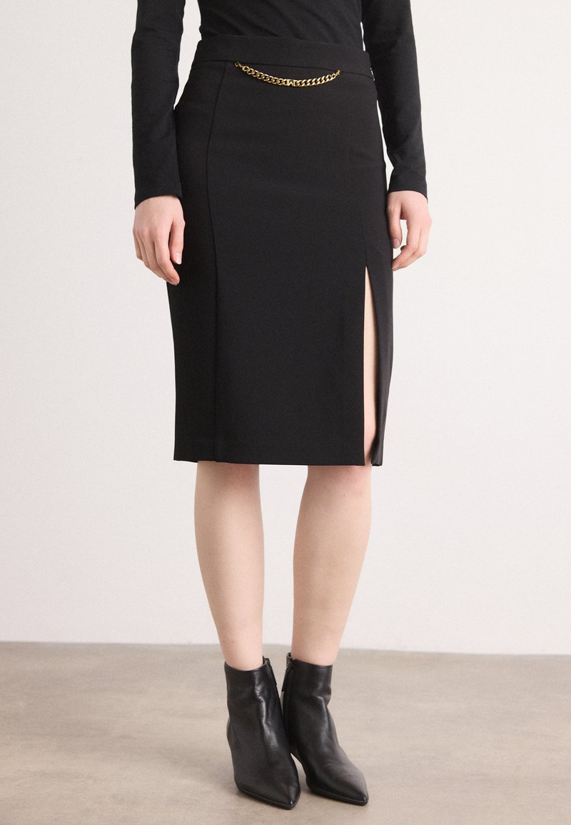 Jupe crayon noire avec une haute fente et un détail en chaîne dorée à la taille. Texture lisse, design ajusté, accompagnée de bottines noires.
