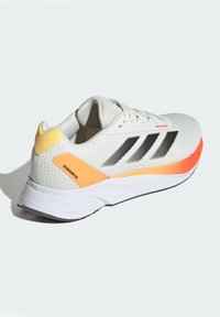 Białe siatkowe buty sportowe z czarnymi paskami, żółtymi i pomarańczowymi akcentami, amortyzowaną podeszwą oraz teksturowaną powierzchnią dla lepszej wentylacji.