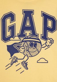 Gul t-skjorte med fete marineblå bokstaver "GAP" og en tegneserie-basketballfigur som har på seg en kappe, løper med joggesko og smiler.