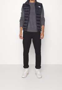 Mężczyzna w czarnej kamizelce puchowej nosi szary sweter, czarne podwinięte spodnie, białe skarpetki i białe trampki, stoi na tle białym.