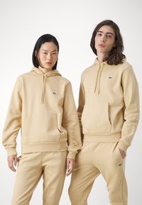 Sudaderas beige con bolsillos tipo canguro, puños de canalé y logotipo verde bordado en el lado izquierdo del pecho, combinadas con pantalones a juego. Textura de tela suave.