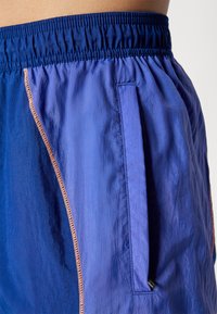 Blaue Sportshorts mit elastischem Bund, ausgestattet mit einer Seitentasche und kontrastierendem orangefarbenem Nähen entlang der Nähte. Leichter, strukturierter Stoff.
