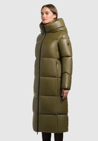 Lange olijfgroene pufferjack met een hoge kraag, glanzende afwerking, verticale naden en met rits afgesloten zakken. Heeft een ritssluiting aan de voorkant en brede mouwen.