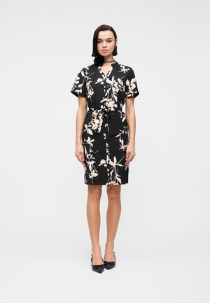 Mujer de pie con un vestido negro hasta la rodilla con estampado floral blanco, mangas cortas, cinturón anudado en la cintura, tacones negros y un accesorio negro en el cuello.