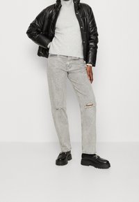 Veste matelassée noire, pull en tricot gris clair, jean gris clair délavé, chaussures à plateforme noires épaisses. Fond uni.