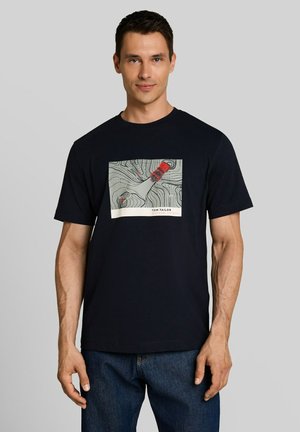 Man wearing a black t-shirt with a red car graphic on a topographic map design, paired with blue jeans, standing against a gray background.