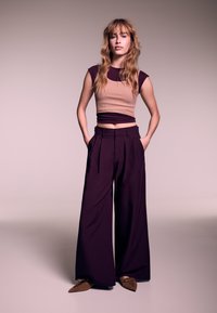 Top corto beige a coste aderente con accenti viola scuro, abbinato a pantaloni bordeaux a gamba larga. Ballerine marroni con punta aggiungono dettaglio.