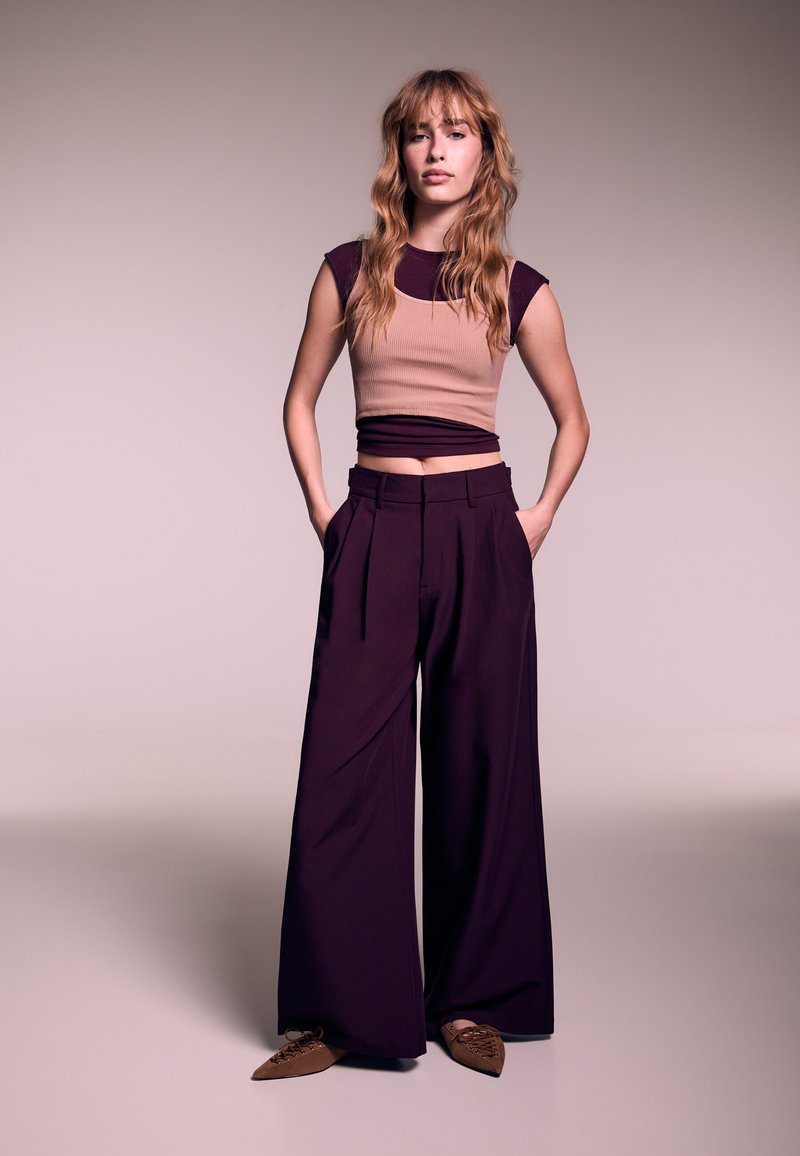 Top corto beige a coste aderente con accenti viola scuro, abbinato a pantaloni bordeaux a gamba larga. Ballerine marroni con punta aggiungono dettaglio.