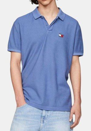 Mężczyzna ubrany w niebieską koszulkę polo z krótkim rękawem, z małym czerwono-biało-niebieskim logo Tommy Hilfiger na piersi, oraz w jasnoniebieskie dżinsy.