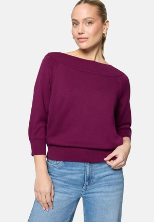 Femme portant un pull violet à épaules dénudées avec des manches trois-quarts et un jean bleu clair, debout devant un fond uni.