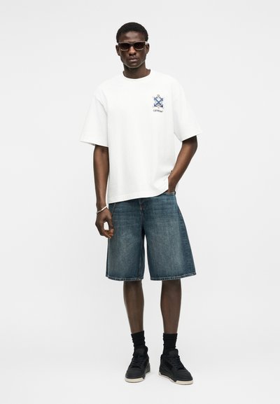 Uomo che indossa occhiali da sole, t-shirt bianca con grafica, pantaloncini di jeans, calzini neri e scarpe da ginnastica nere, in piedi con una mano in tasca.