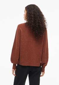 Hosszú göndör hajú nő, rozsda színű kötött pulóverben, amelynek puffos ujja van, és fekete nadrágot visel, hátulról nézve, fehér háttér előtt.