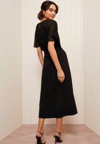 Abito midi nero con maniche corte trasparenti e leggere, vita aderente e dettaglio a fessura sulla schiena. Abbinato a tacchi alti con cinturino.