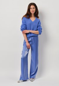 Blaues Wollgemischpullover mit V-Ausschnitt und übergroßen Ärmeln, kombiniert mit glänzenden blauen Satinhosen und silbernen spitzen Schuhen.