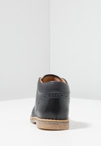 Scarpa di pelle nera con una texture liscia, caratterizzata da dettagli brogue sul tallone e una suola marrone chiaro. Design minimalista.