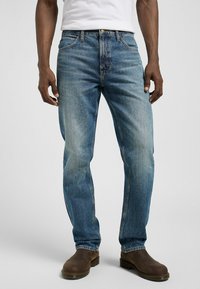 Jeans de mezclilla azul con corte recto, que presentan un lavado descolorido, diseño de cinco bolsillos y herrajes metálicos. Llevados con botas de cuero marrón.