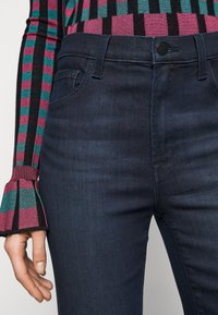 Vaqueros de mezclilla de color azul oscuro con una textura suave, que cuentan con un bolsillo frontal, trabillas para el cinturón y un cierre de botón. Puños de suéter a rayas en rosa y verde azulado.