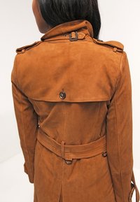 Brązowy zamszowy płaszcz trench z paskiem w talii, zapięciem na guziki z tyłu oraz pagonami na ramionach. Gładka tekstura z dopasowaną sylwetką.