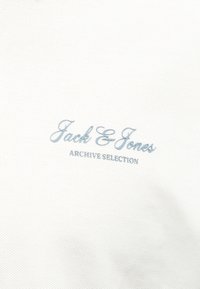 Λευκό υφασμάτινο ύφασμα με υφασμάτινη υφή και κεντημένο κείμενο σε ανοιχτό μπλε χρώμα που γράφει "Jack & Jones ARCHIVE SELECTION" στο κέντρο.