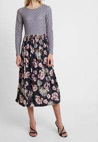 Vrouw in een lange jurk met een klein geometrisch patroon aan de bovenkant en een marineblauwe bloemenrok, gecombineerd met zwarte hoge hakken.