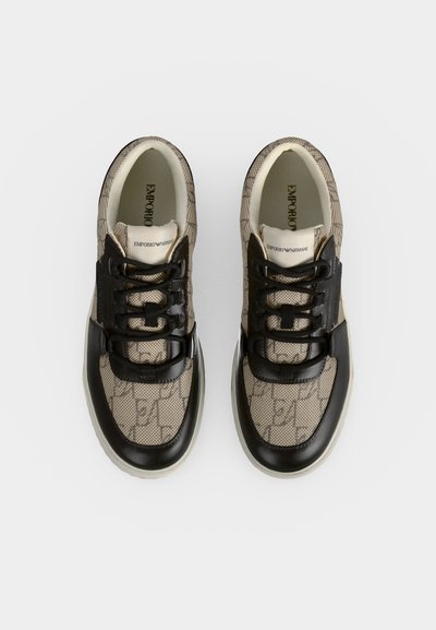 Ζευγάρι μπεζ και μαύρα σχεδιαστικά sneakers με μοτίβο λογότυπου και μαύρα κορδόνια τοποθετημένα δίπλα δίπλα σε λευκό φόντο.