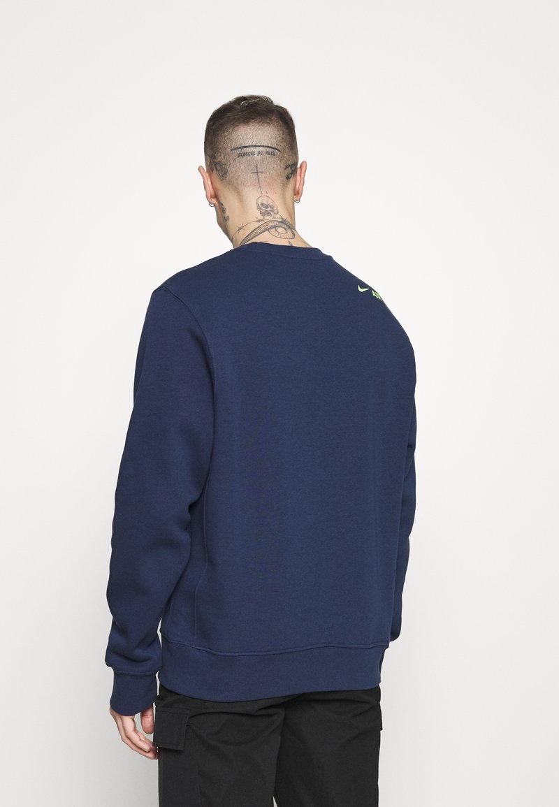 Uomo con tatuaggi sulla parte posteriore della testa e del collo, che indossa una felpa blu navy e pantaloni cargo neri, di spalle alla fotocamera.