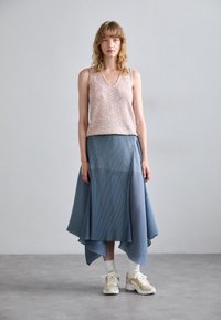 Beige paljetteret tanktop kombinert med en flytende, plissert blå skjørt. Modellen bruker chunky hvite sneakers. Enkel bakgrunn, nøytrale toner.