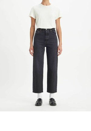 Personne portant une chemise blanche à manches courtes, un jean noir taille haute à jambes droites, des chaussettes blanches et des mocassins noirs, debout devant un fond blanc.