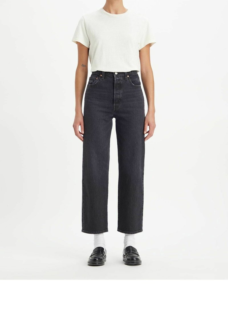 Personne portant une chemise blanche à manches courtes, un jean noir taille haute à jambes droites, des chaussettes blanches et des mocassins noirs, debout devant un fond blanc.