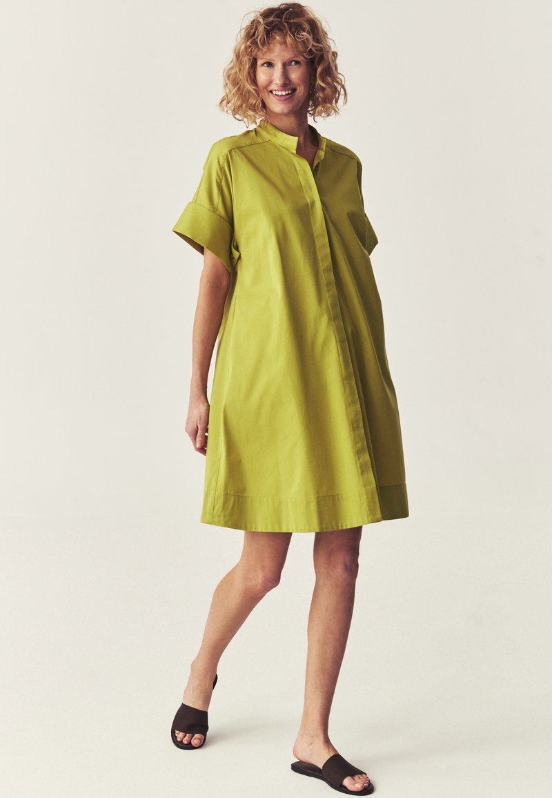 Femme souriante aux cheveux bouclés porte une robe ample vert vif à manches courtes et des sandales noires slides, marchant sur un fond clair uni.