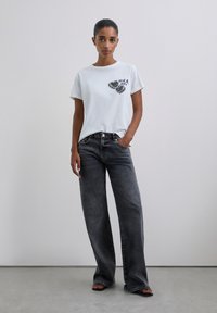 Kobieta opierająca się o gładką ścianę, ubrana w biały t-shirt z grafiką, wyblakłe czarne dżinsy w szerokim kroju i czarne klapki, z jedną ręką w kieszeni.