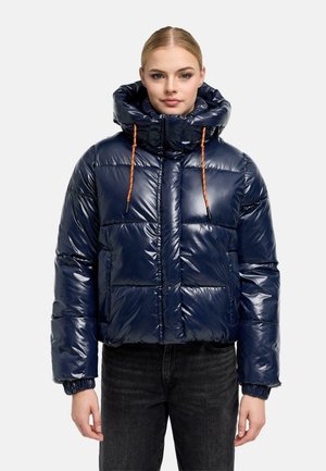 Jeune femme portant une doudoune bleu marine brillante avec un col montant et des cordons orange, associée à un jean noir, debout devant un fond blanc.