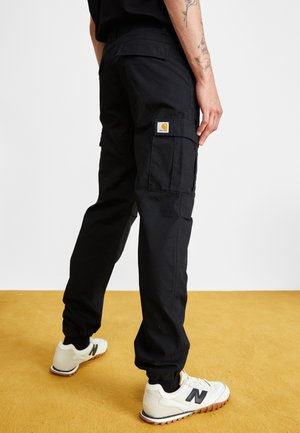 Person trägt schwarze Cargohose mit Logopatch an der Seitentasche und weiße Sneaker mit schwarzem "N"-Logo, steht auf einem senfgelben Boden.