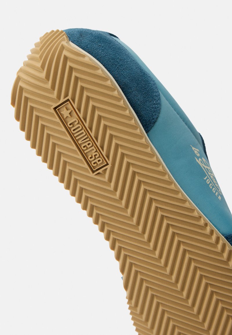 Blauer Converse Slip-On-Sneaker mit strukturierter, bräunlicher Sohle, auf der obereren Seite mit dem "Converse"-Logo und dem Text "le coq sportif JOGGER" versehen.